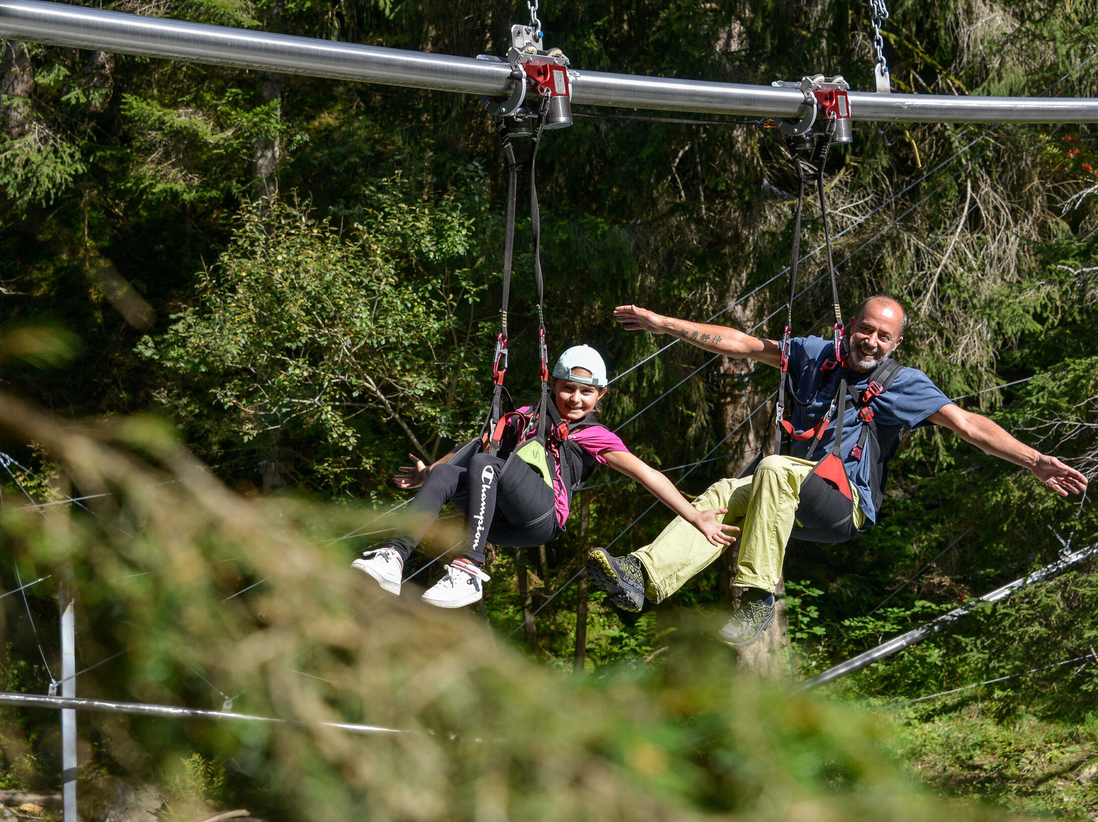 Zwei Personen in Sicherheitsausrüstung gleiten an einer Fly-Line-GmbH-Seilbahn durch einen grünen Wald, der Almberg-Erlebnis-Hintergrund vermittelt eine abenteuerliche Outdoor-Atmosphäre.