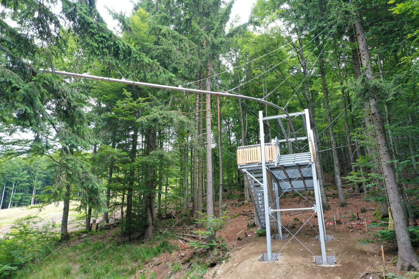 Almberg Fly-Line Baukonstruktion im Wald: Stahlrahmen, Holzplattform und Geländer, grüne Nadelbäume, Bauarbeiten sichtbar, Baumreihen im Hintergrund