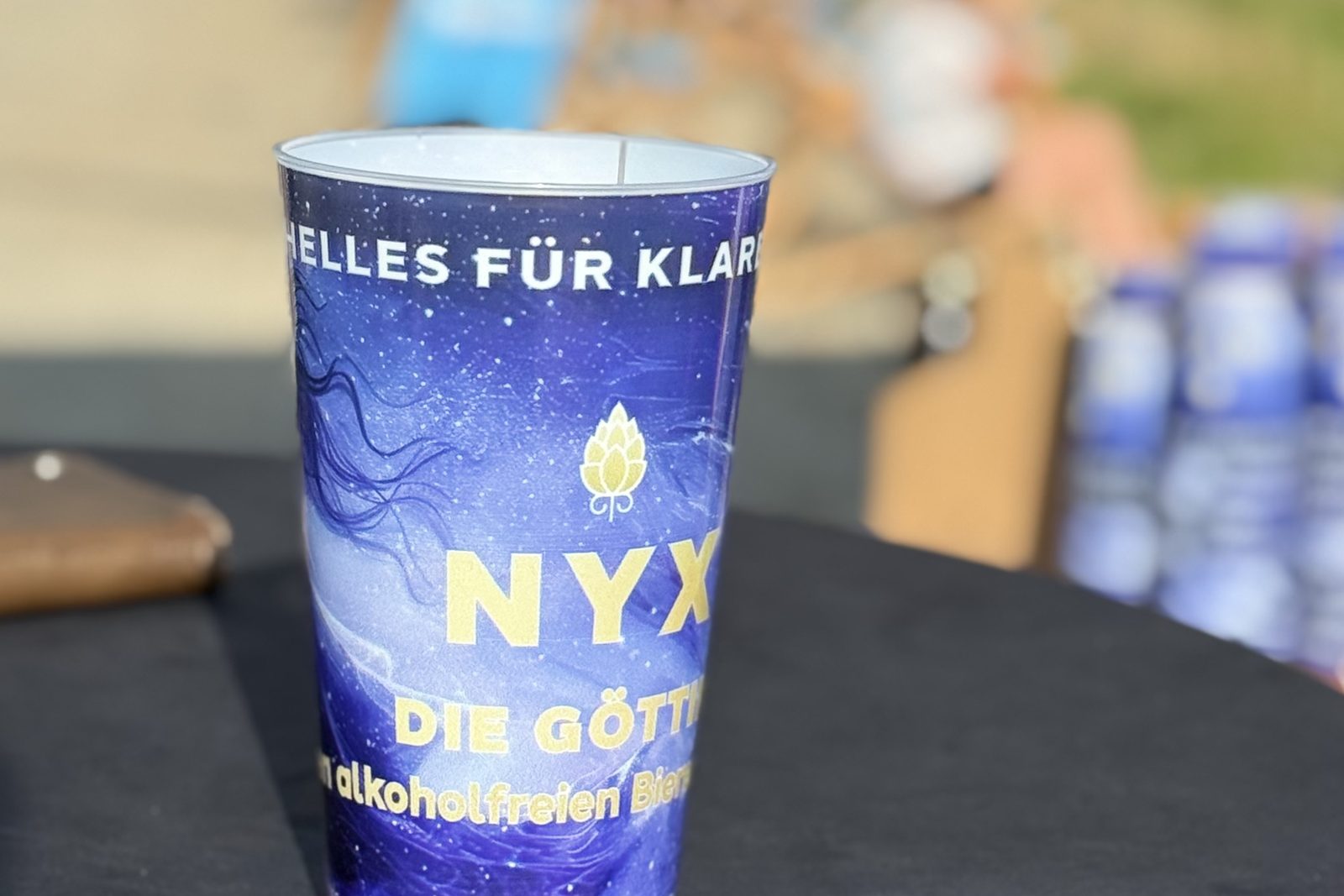 Ein blauer NYX-Becher mit dem Aufdruck „Die Göttin“ steht auf einem Tisch; er enthält das alkoholfreie Bier von Kanzel Bräu.