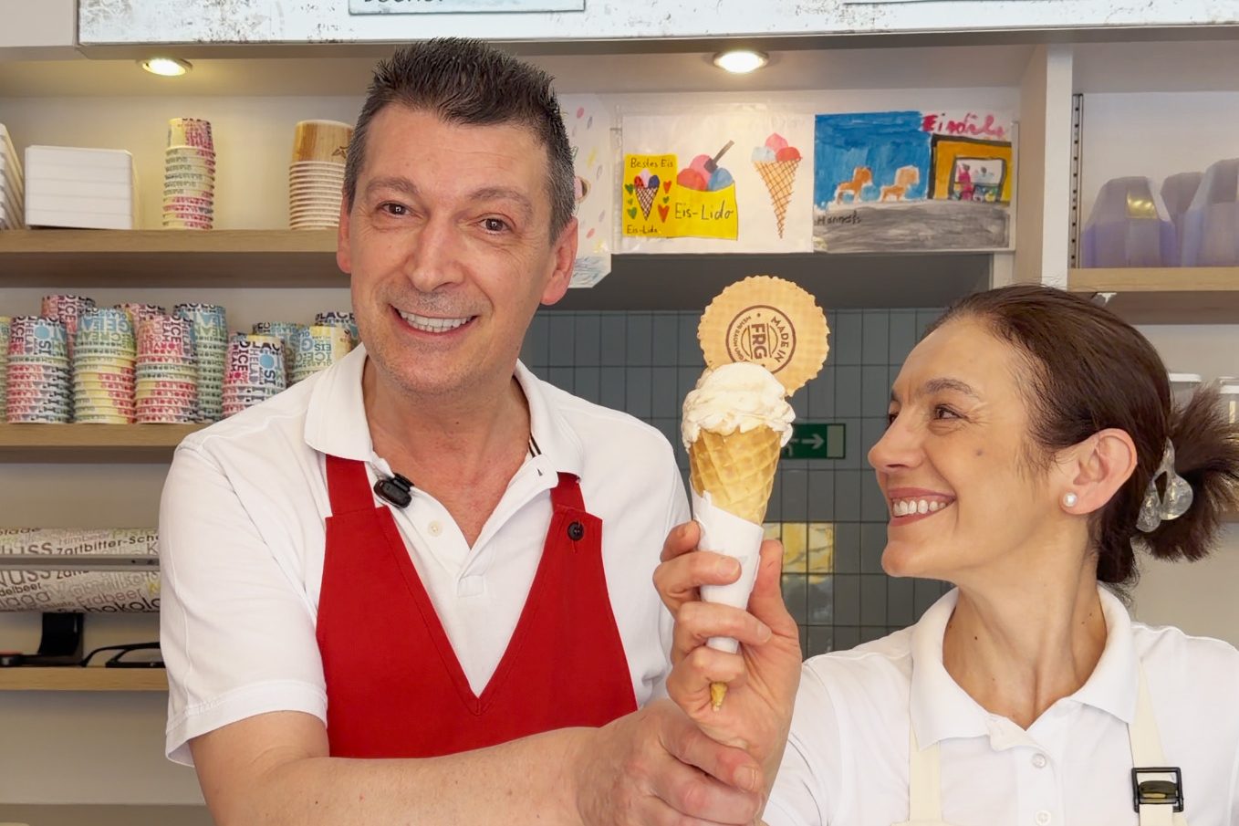 Bei Eis Lido in Freyung hält ein Mitarbeiter lächelnd eine Waffel mit weißem Eis, während eine Kollegin ihn anlächelt.