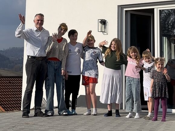 Steffi und Martin Dorsch stehen mit ihren sieben Kindern vor ihrem neuen Zuhause im Landkreis Freyung-Grafenau und winken.