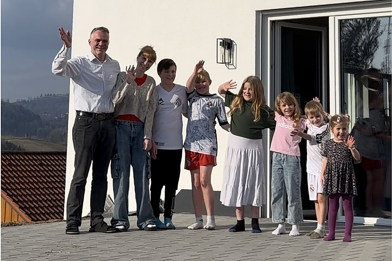 Steffi und Martin Dorsch stehen mit ihren sieben Kindern vor ihrem neuen Zuhause im Landkreis Freyung-Grafenau und winken.