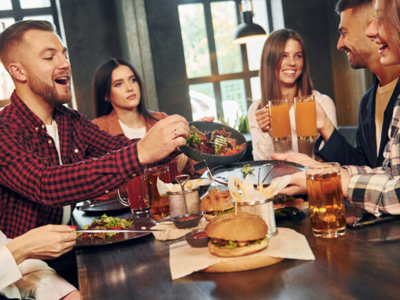 Freunde teilen Burger und Salat an einem Restauranttisch und stoßen mit Getränken an, während sie gemeinsam essen und lachen.