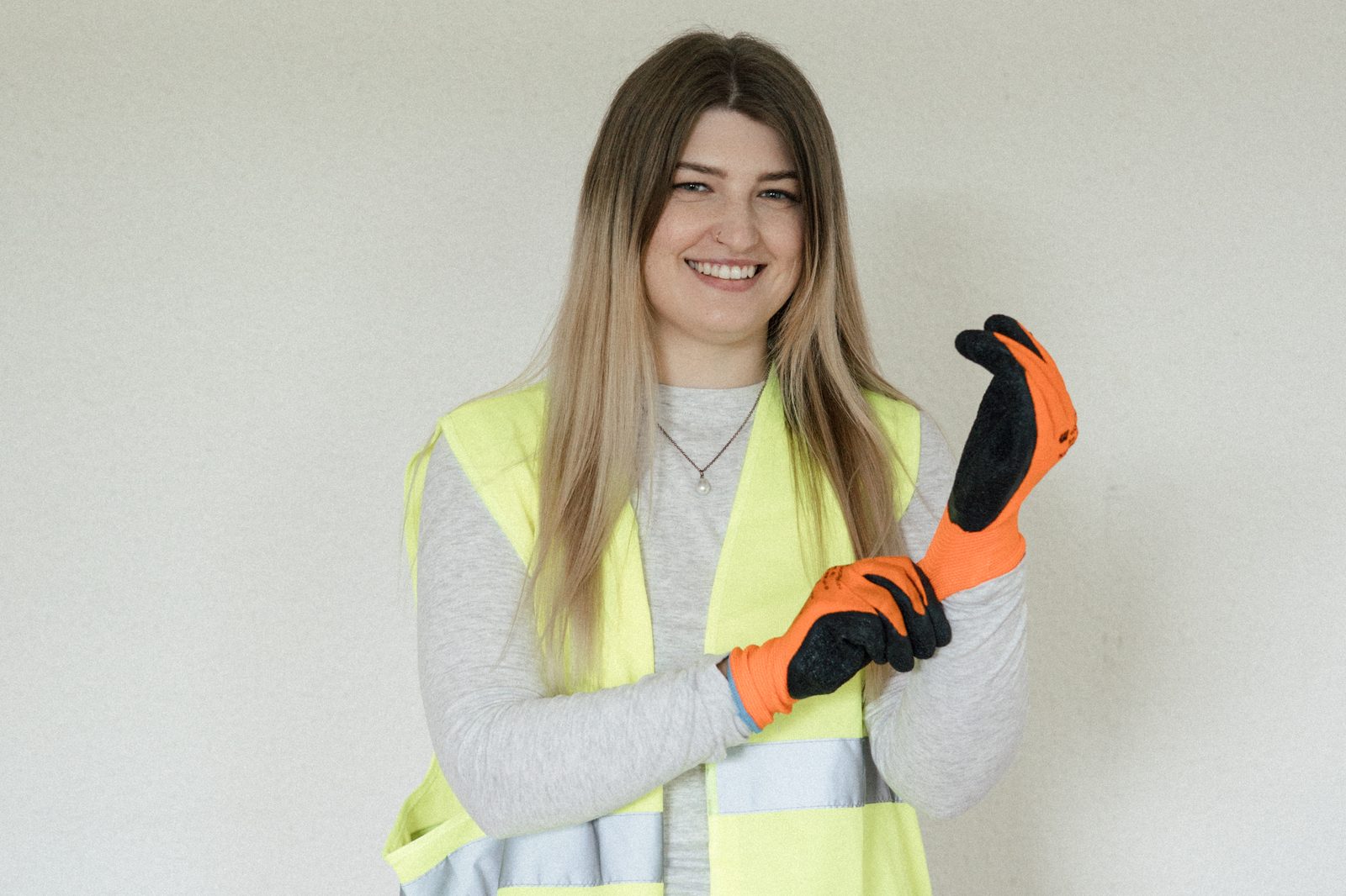 Eine lächelnde Jobtesterin zieht orange-schwarze Arbeitshandschuhe an und trägt eine gelbe Warnweste beim Ausprobieren eines Berufs.