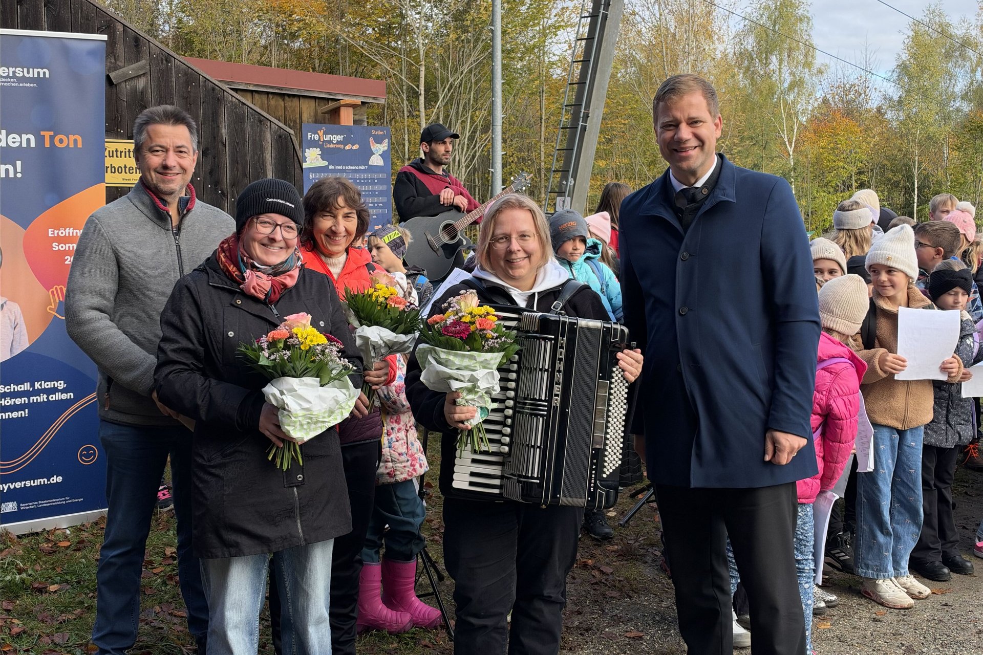 Beteiligte mit Blumen und eine Akkordeonspielerin feiern die Eröffnung des Freyunger Liederwegs mit Kindern am Geyersberg.