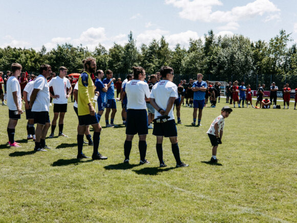 Auf dem Sportgelände des TV Freyung stehen Spieler in weißen und blauen Trikots beim Firmen-Fußballturnier auf dem Rasen.