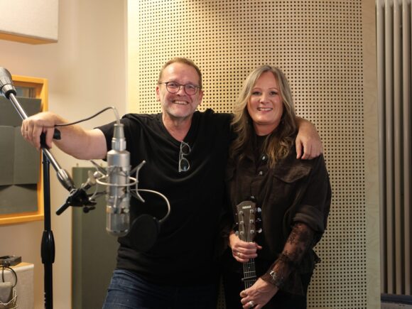 Im Tonstudio lächeln Marion Bauer und ein Mann nebeneinander; vor ihnen stehen Mikrofone, und Bauer hält ihre Yamaha-Gitarre.