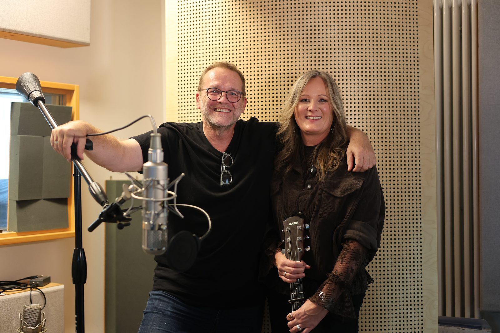 Im Tonstudio lächeln Marion Bauer und ein Mann nebeneinander; vor ihnen stehen Mikrofone, und Bauer hält ihre Yamaha-Gitarre.