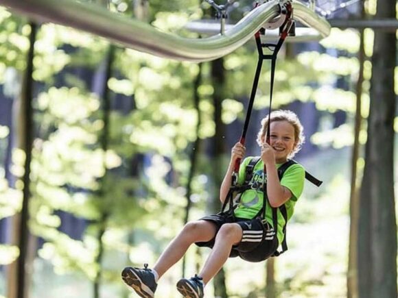 Ein Junge fährt lächelnd auf der Fly-Line in Mitterdorf an einer Schiene durch den Wald und hält sich an den Gurten fest.