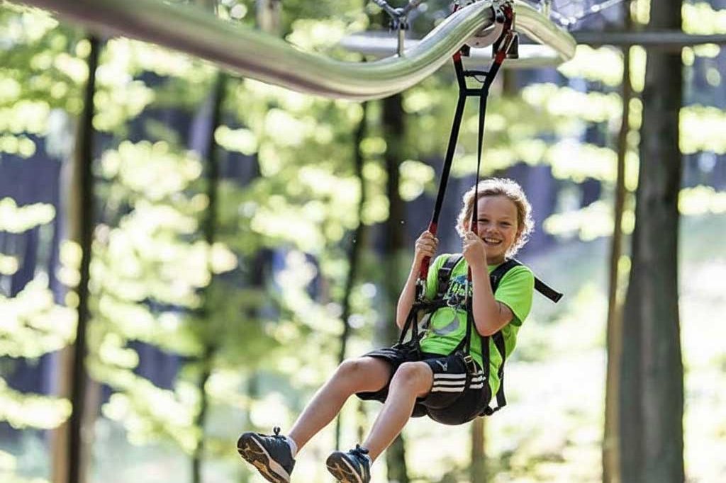 Ein Junge fährt lächelnd auf der Fly-Line in Mitterdorf an einer Schiene durch den Wald und hält sich an den Gurten fest.