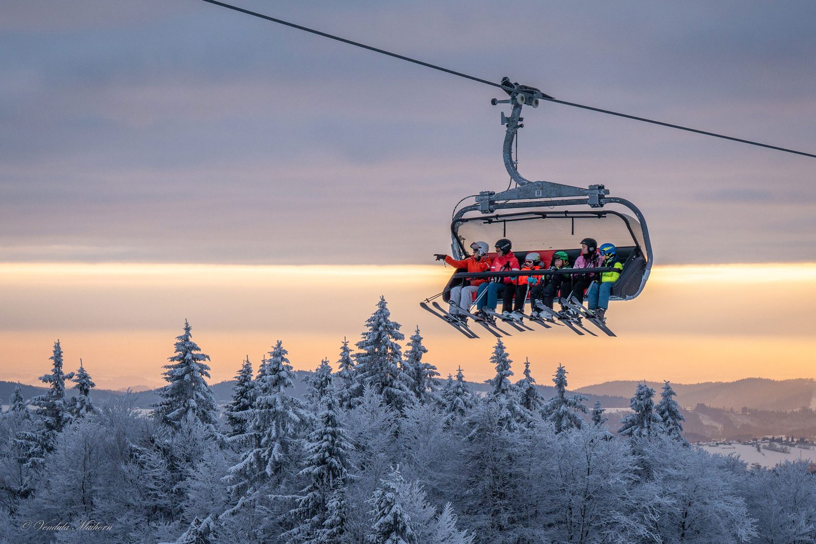 Mehrere Skifahrer fahren mit einer Sesselbahn über verschneite Baumwipfel in Mitterdorf; die Wintersonne leuchtet am Horizont.