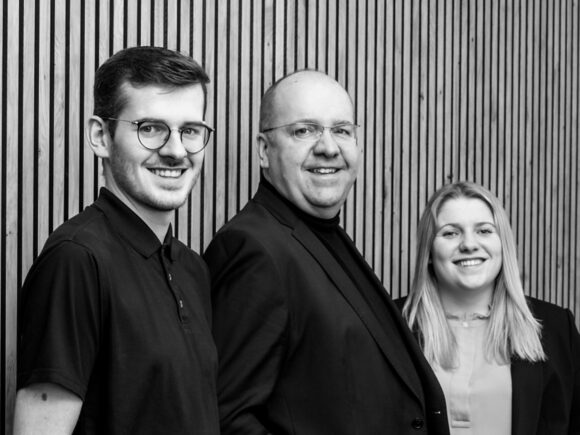 Drei lächelnde Mitglieder des SCHRAML.IT-Teams stehen vor einer Holzleistenwand; der IT-Dienstleister feiert 30 Jahre in Freyung.